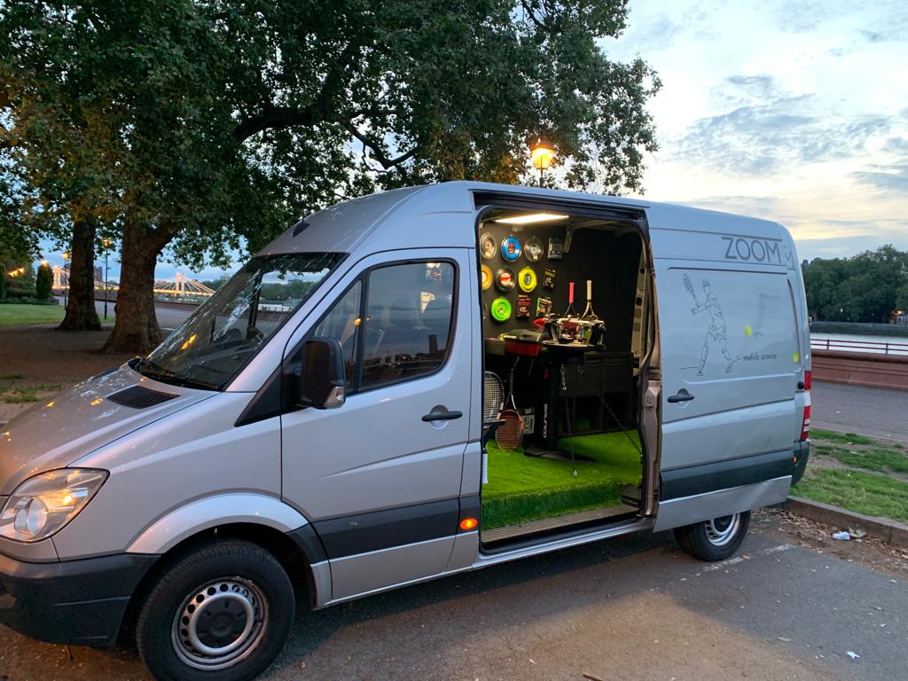 ZOOM Stringing mobile van parked by the river at dusk, side door open showing the stringing machine and rackets inside